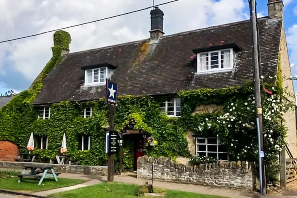 A traditional stone building covered in green ivy with a steep, dark roof and white-framed windows. There is a wooden signpost with a blue star outside, a stone wall along the front, and picnic tables with umbrellas on the grass. The sky is partly cloudy. A traditional stone building covered in green ivy with a steep, dark roof and white-framed windows. There is a wooden signpost with a blue star outside, a stone wall along the front, and picnic tables with umbrellas on the grass. The sky is partly cloudy.