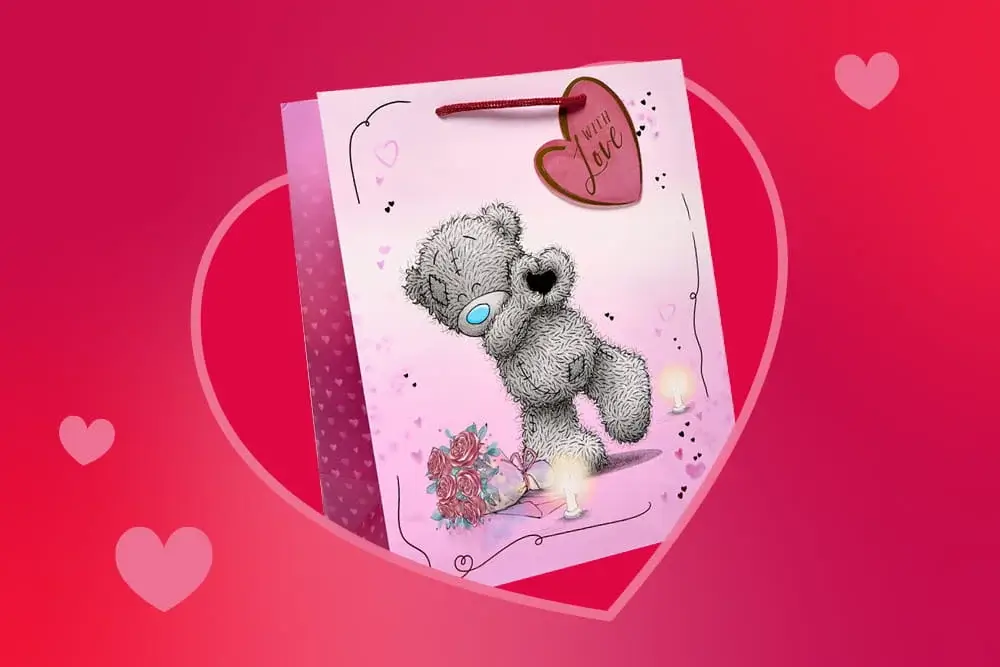 Pink gift bag featuring a cute grey teddy bear holding a heart shape, surrounded by soft pink accents and floral details.