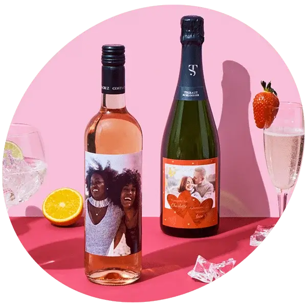 Two personalised wine bottles on a pink background, one rosé and one sparkling, each with custom photo labels. Surrounding items include a glass with an orange slice, a champagne flute with a strawberry, and scattered ice cubes.