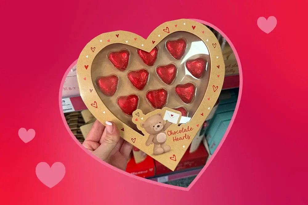 Heart‑shaped box of red foil chocolate hearts with a cute teddy bear illustration, held in front of a store display.