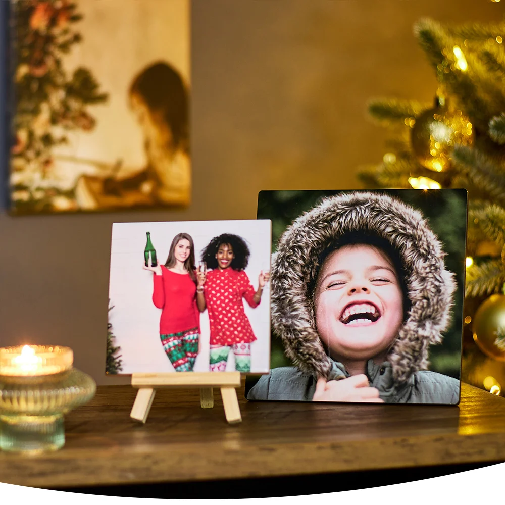 Cozy Christmas scene with two framed photos on a wooden table—one of two people in festive outfits holding drinks, and another of a person laughing in a winter coat, with blurred decorations and a lit candle in the background.
