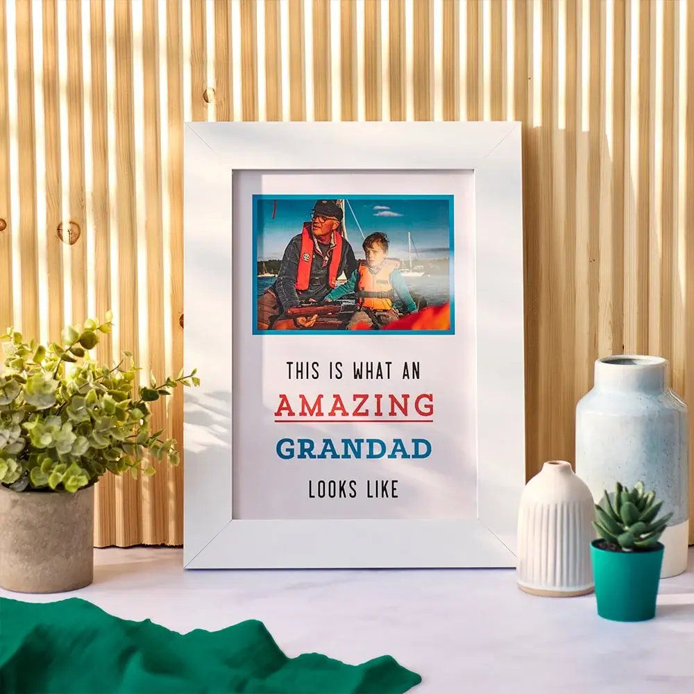 A framed photo of a man and a child with the text 'THIS IS WHAT AN AMAZING GRANDAD LOOKS LIKE' displayed below the photo. The frame is placed on a table with various decorative items, including potted plants and vases, against a wooden slat background.