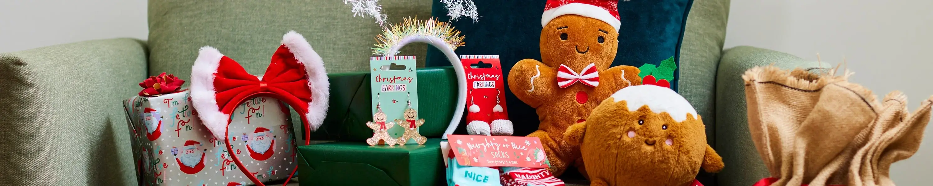A festive display on a green armchair featuring Christmas-themed items, including wrapped gifts, a headband with red and white ears, gingerbread man earrings, a plush gingerbread toy wearing a Santa hat, a round Christmas pudding plush, and a burlap sack. Snowflake decorations hang above the arrangement.