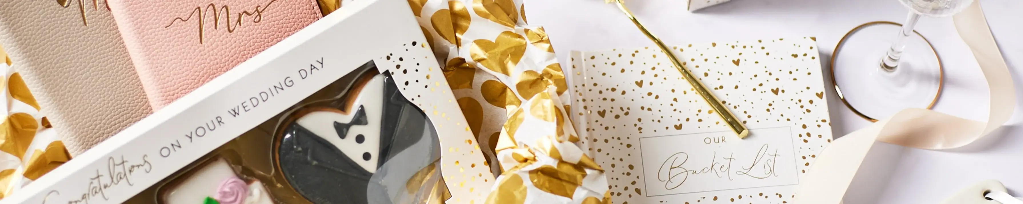 'A wedding-themed flat lay image featuring a box of cookies, two pink and beige notebooks labeled 'Mrs', a white and gold polka-dotted gift box with the text 'Our Bucket List', and a champagne glass. The box of cookies has the text 'Congratulations on your wedding day' on it.' 'A wedding-themed flat lay image featuring a box of cookies, two pink and beige notebooks labeled 'Mrs', a white and gold polka-dotted gift box with the text 'Our Bucket List', and a champagne glass. The box of cookies has the text 'Congratulations on your wedding day' on it.'