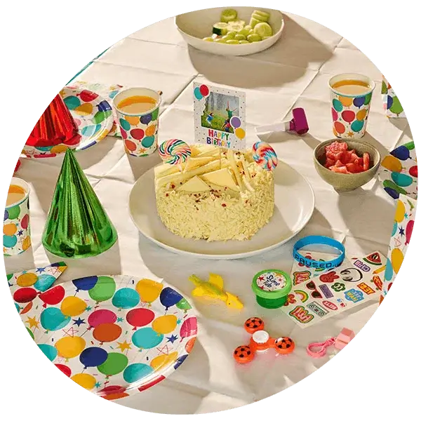 Colourful birthday party table with a birthday cake, party hats, balloon-print tableware, drinks, sweets, and children's party favours.