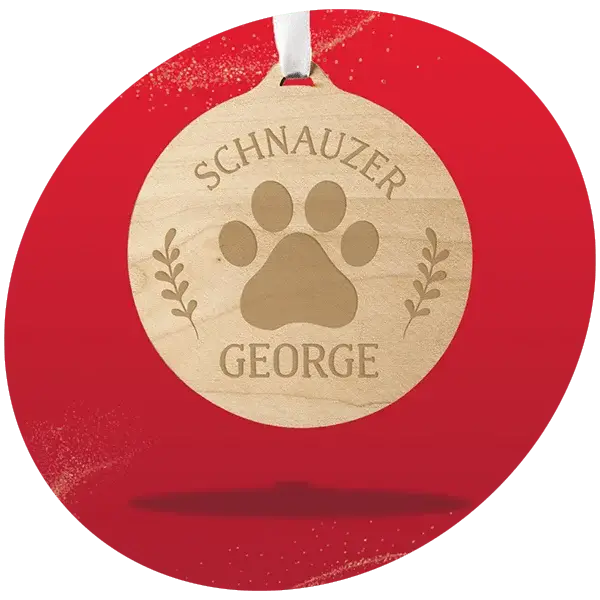Round wooden Christmas ornament engraved with a paw print design and the text Schnauzer George, decorated with small leaf motifs and hanging from a white ribbon, displayed against a bright red background.