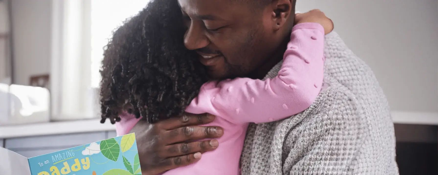 A person with short curly hair wearing a pink long-sleeve shirt is hugging another person wearing a gray sweater. The face of the person being hugged is partially obscured. In the foreground, there is a colorful card with the text 'to an amazing daddy' and illustrations of leaves. A person with short curly hair wearing a pink long-sleeve shirt is hugging another person wearing a gray sweater. The face of the person being hugged is partially obscured. In the foreground, there is a colorful card with the text 'to an amazing daddy' and illustrations of leaves.