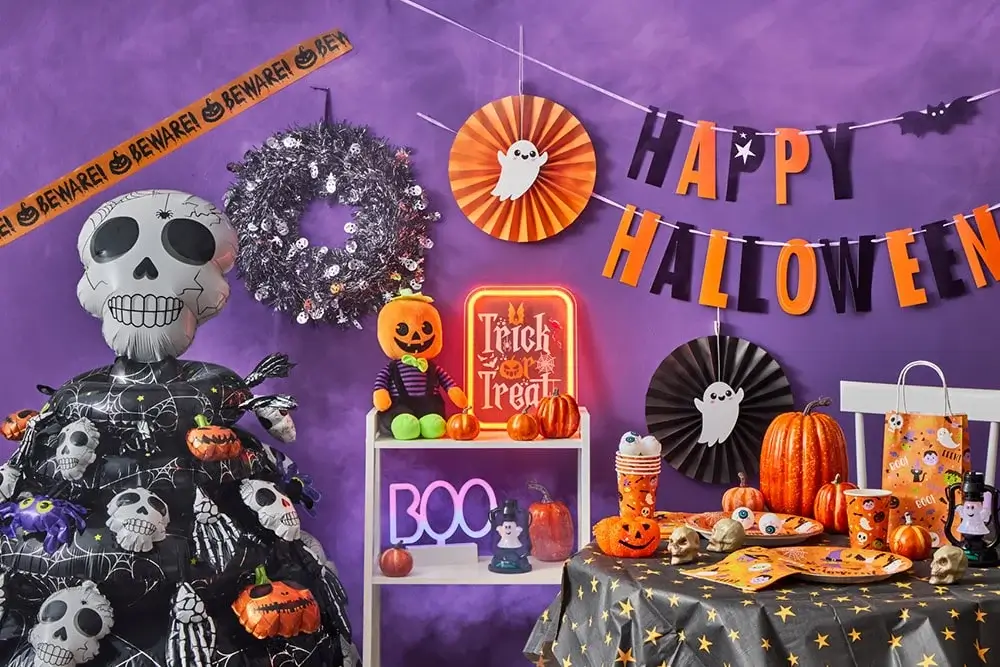 Halloween display with an inflatable skeleton, caution tape, silver wreath, pumpkins, Trick or Treat sign, and Happy Halloween banner with ghost cutouts on a purple background.