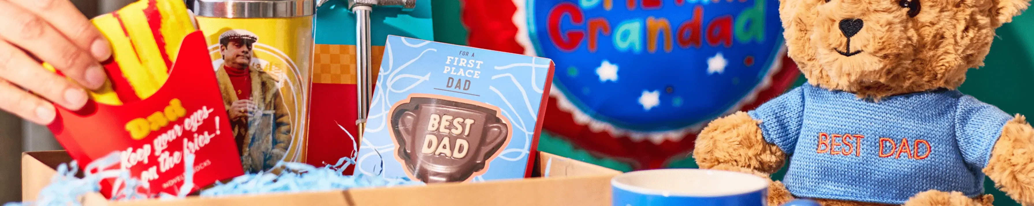 A close-up image of a gift box containing various items for Father's Day. The box includes a red container with yellow French fries labeled 'Best Dad', a blue card with the text 'First Place Dad' and an image of a trophy that says 'Best Dad', and a teddy bear wearing a blue sweater with the words 'Best Dad' embroidered on it. There is also some shredded blue paper filling in the box.' A close-up image of a gift box containing various items for Father's Day. The box includes a red container with yellow French fries labeled 'Best Dad', a blue card with the text 'First Place Dad' and an image of a trophy that says 'Best Dad', and a teddy bear wearing a blue sweater with the words 'Best Dad' embroidered on it. There is also some shredded blue paper filling in the box.'