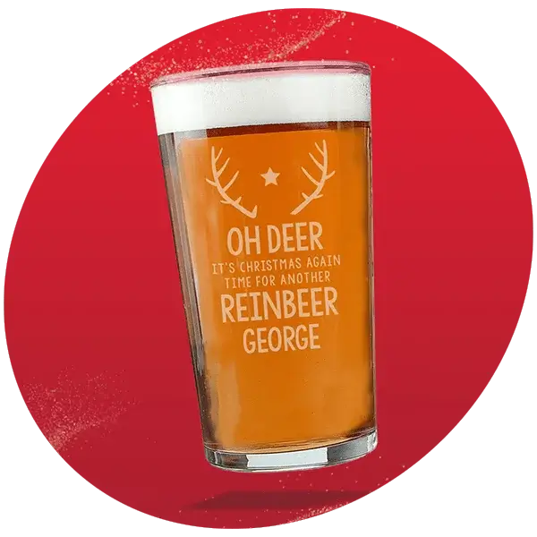 Pint glass filled with beer, decorated with antlers and a star, featuring the text Oh Deer It's Christmas Again Time For Another Reinbeer George, set against a vibrant red background with white speckles.