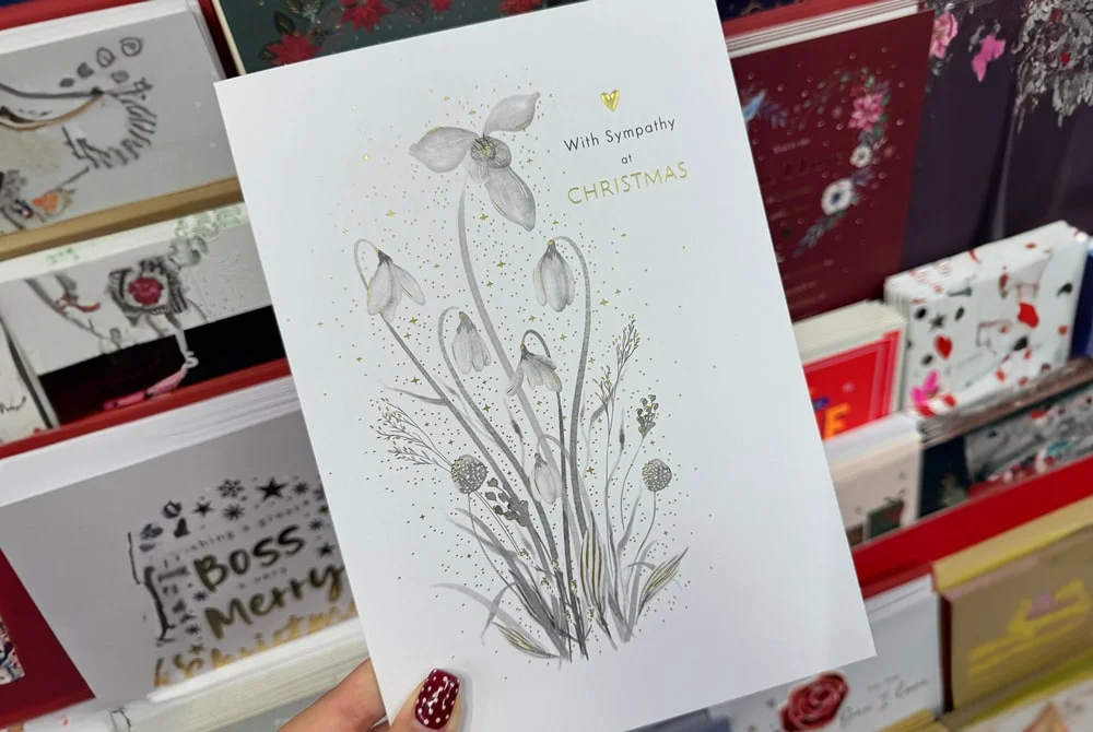 A hand holding a white sympathy Christmas card with an illustration of delicate flowers and gold accents, displayed among other greeting cards in a store. A hand holding a white sympathy Christmas card with an illustration of delicate flowers and gold accents, displayed among other greeting cards in a store.