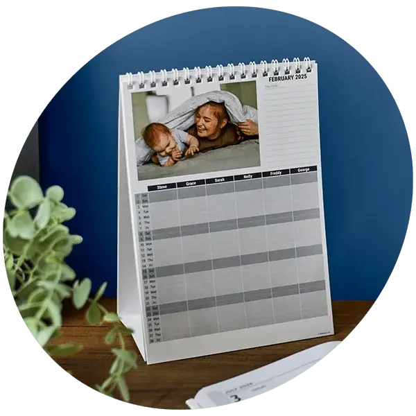 A spiral-bound desk calendar open to February 2025, featuring a photo of a baby lying under a blanket with an adult. The calendar includes a grid layout for organizing names and schedules, placed on a wooden surface with a plant partially visible in the foreground and a blue wall in the background.