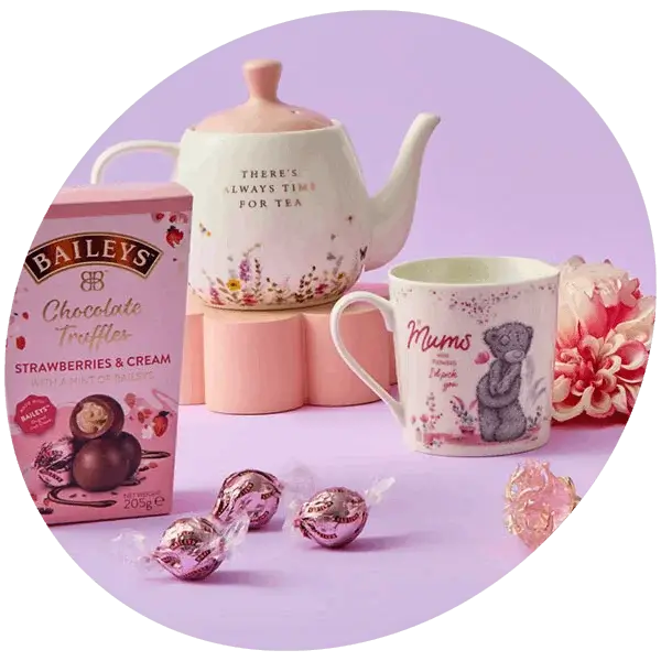A pastel tea‑themed gift set featuring a floral teapot, a matching Mum mug with a teddy bear design, Baileys chocolate truffles, and wrapped sweets arranged on a lilac surface.