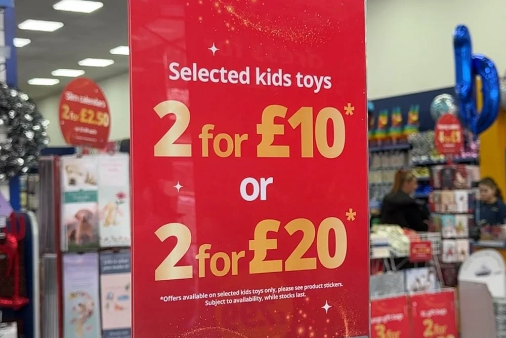 A red promotional sign in a store displaying an offer for selected kids’ toys, reading “2 for £10 or 2 for £20,” with additional details about availability and conditions at the bottom. A red promotional sign in a store displaying an offer for selected kids’ toys, reading “2 for £10 or 2 for £20,” with additional details about availability and conditions at the bottom.