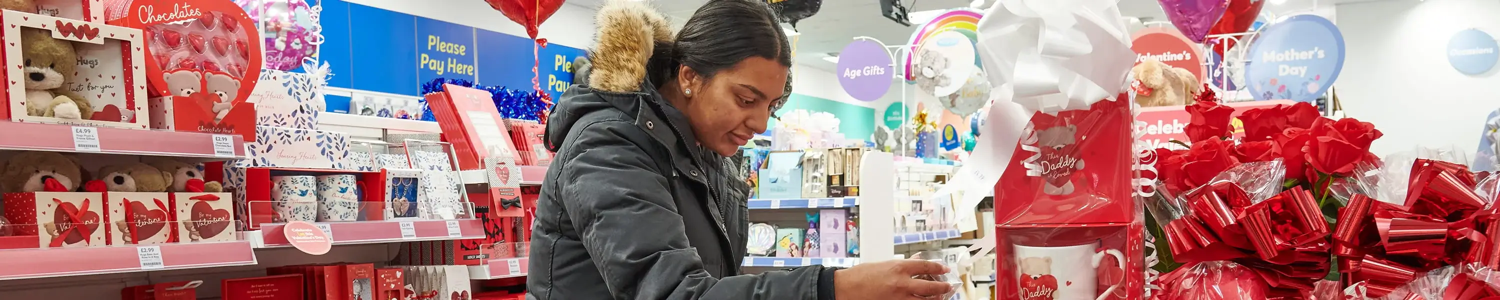 Shopper browsing a store aisle filled with Valentine’s Day gifts, including red roses, balloons, mugs, and heart‑themed decorations.