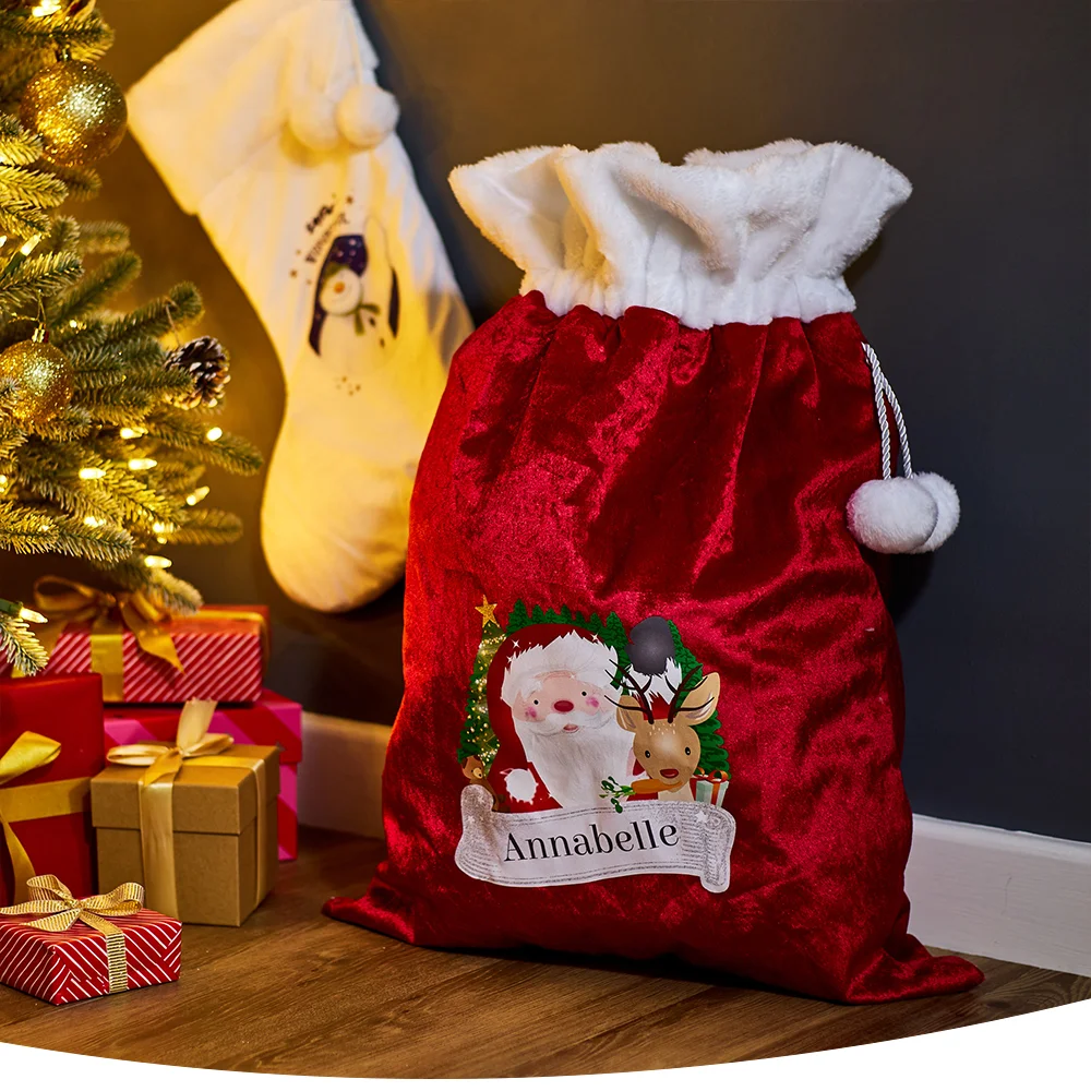 Christmas scene with a red velvet gift bag labeled 'Annabelle' featuring Santa and a reindeer, next to wrapped presents and a decorated tree, with a penguin-themed stocking hanging in the background.
