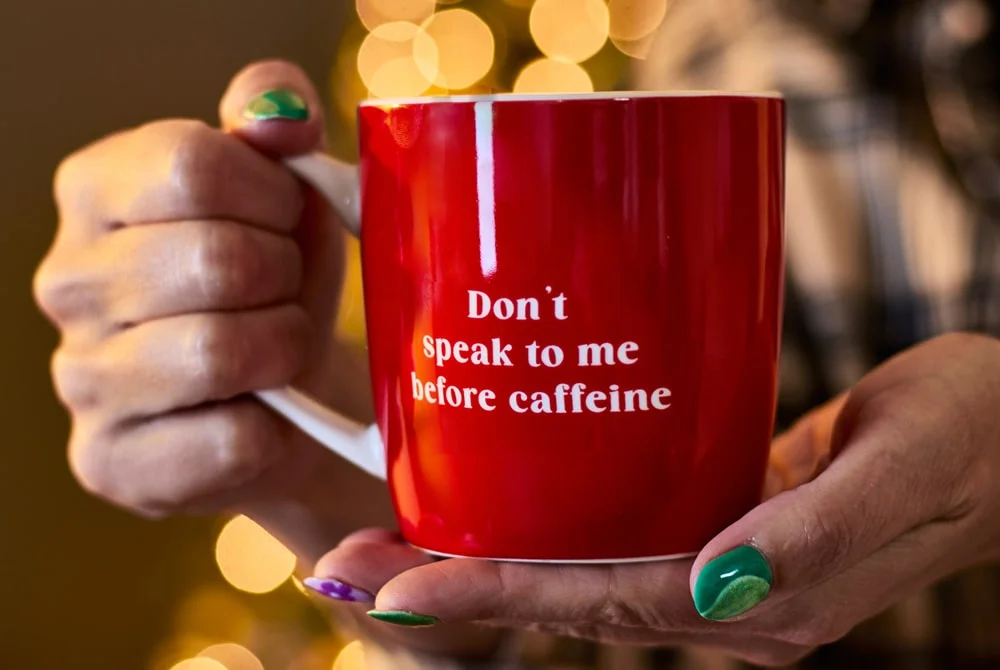 A person holding a red coffee mug with white text that reads “Don’t speak to me before caffeine,” against a blurred background of warm golden lights. A person holding a red coffee mug with white text that reads “Don’t speak to me before caffeine,” against a blurred background of warm golden lights.