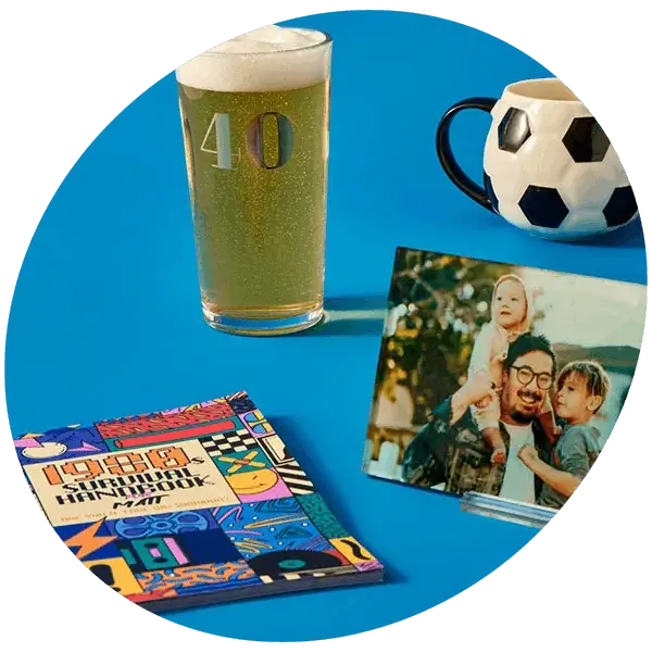 A display of birthday gift items on a blue surface, including a beer glass marked 40, a football‑themed mug, a colourful book, and a photo in a clear stand.