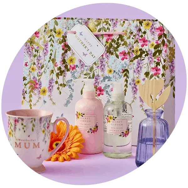 A floral Mother’s Day gift set featuring a pastel flower‑patterned gift bag, a matching mug, floral‑scented lotions, and a blue reed diffuser bottle arranged with an orange flower on a lilac background.