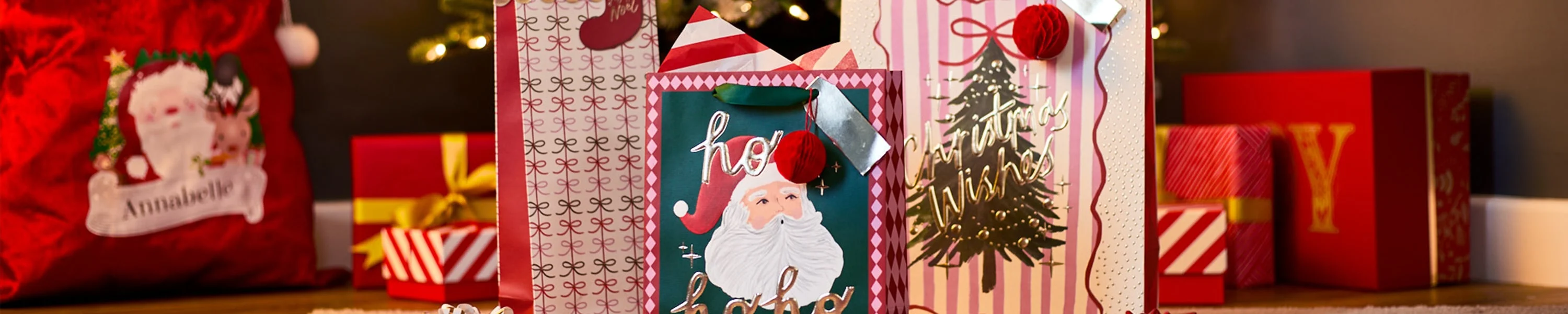 Festive gift bags and wrapped presents with Christmas designs, including Santa and a decorated tree, arranged under a Christmas tree with warm lights in the background. Festive gift bags and wrapped presents with Christmas designs, including Santa and a decorated tree, arranged under a Christmas tree with warm lights in the background.