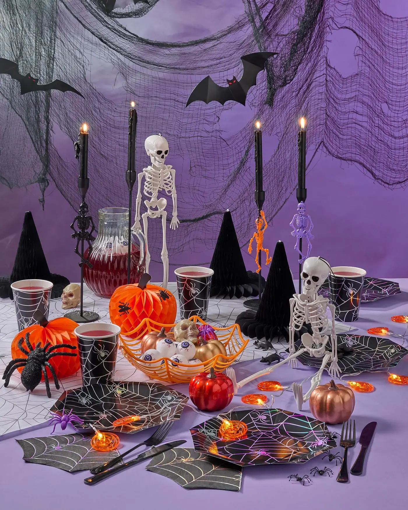 Halloween-themed table with skeletons, pumpkins, spiderweb plates, and spooky decorations against a purple backdrop. Halloween-themed table with skeletons, pumpkins, spiderweb plates, and spooky decorations against a purple backdrop.
