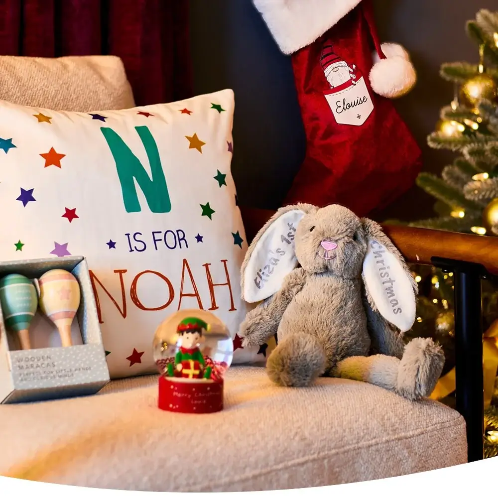 Cozy Christmas scene with personalized pillow, red stocking, plush bunny, maracas, and elf snow globe near a lit Christmas tree.