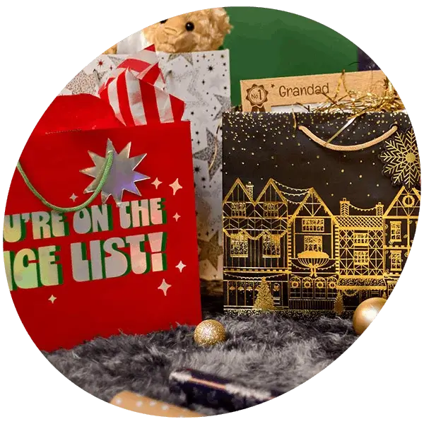 Festive Christmas gift bags filled with gifts and cards, displayed on a soft grey rug with gold bauble decorations.