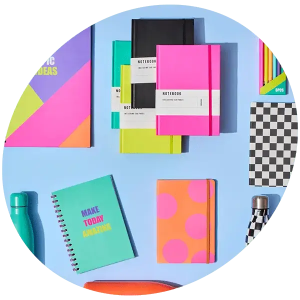 Circular image of colorful stationery on a light blue background, including labeled notebooks, a 'MAKE TODAY AMAZING' spiral notebook, a checkered notebook, colored pencils, and two water bottles.