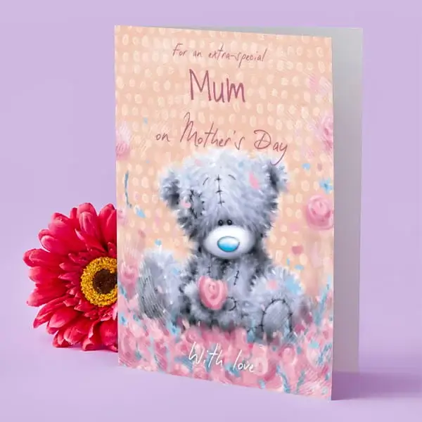 A pastel Mother’s Day card featuring a cute grey teddy bear holding pink flowers, set against a soft peach background with floral accents, displayed next to a bright pink flower on a lilac surface.