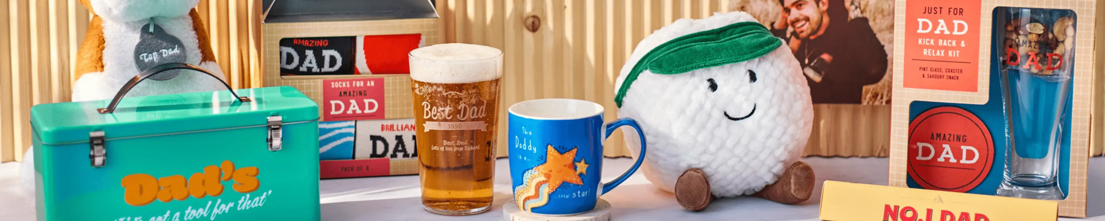 A collection of Father's Day gifts is displayed on a table. Items include a green lunchbox with 'Dad's' written on it, several books titled 'DAD', a beer glass filled with beer, a blue mug with an orange fox design, a plush golf ball toy wearing a green visor, and various cards and small items celebrating dads.' A collection of Father's Day gifts is displayed on a table. Items include a green lunchbox with 'Dad's' written on it, several books titled 'DAD', a beer glass filled with beer, a blue mug with an orange fox design, a plush golf ball toy wearing a green visor, and various cards and small items celebrating dads.'