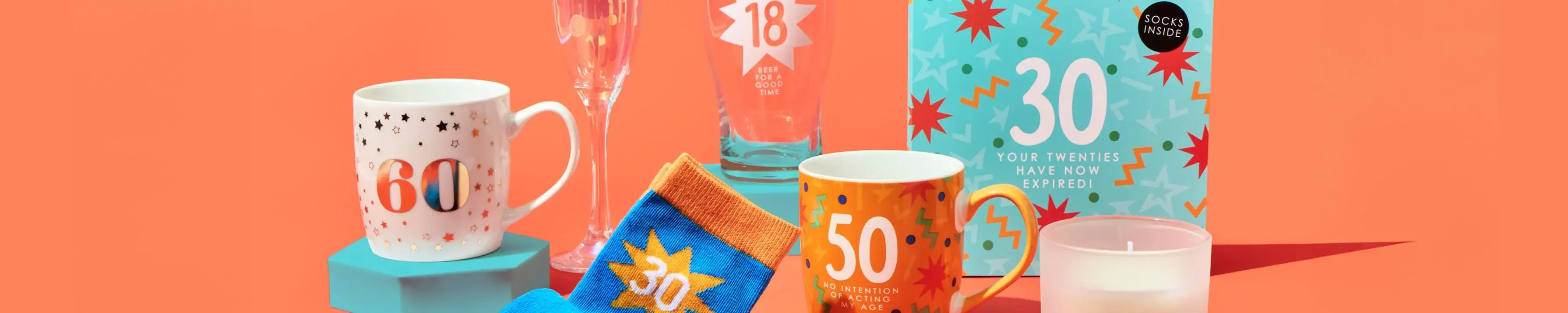 'The image shows a collection of birthday-themed items against an orange background. There is a white mug with the number 60 and star patterns, a champagne flute with the number 18 and the text 'It's time for good times', a pair of blue socks with orange accents and the number 30, an orange mug with the number 50 and the text 'No mention of age required', a blue box with colorful starburst patterns, the number 30, and the text 'Your twenties have now expired! Socks inside', and a white candle in a glass container.' 'The image shows a collection of birthday-themed items against an orange background. There is a white mug with the number 60 and star patterns, a champagne flute with the number 18 and the text 'It's time for good times', a pair of blue socks with orange accents and the number 30, an orange mug with the number 50 and the text 'No mention of age required', a blue box with colorful starburst patterns, the number 30, and the text 'Your twenties have now expired! Socks inside', and a white candle in a glass container.'