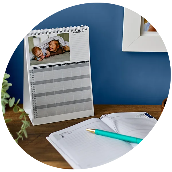 A spiral-bound desk calendar open to February 2025, featuring a photo of a baby lying under a blanket with an adult. The calendar includes a grid layout for organizing names and schedules, placed on a wooden surface with a plant partially visible in the foreground and a blue wall in the background.