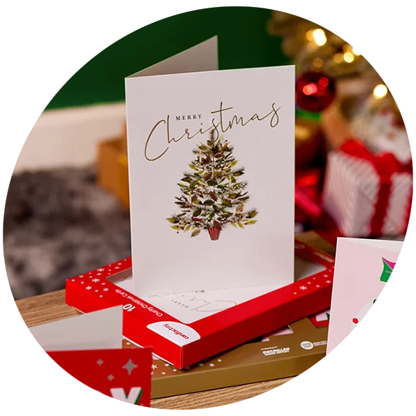 A boxed Christmas card set displayed on a wooden surface, featuring a greeting card with a decorated Christmas tree and gold script text reading ‘Merry Christmas.’ The card stands on top of red and gold card packaging, with wrapped gifts and festive lights softly blurred in the background.