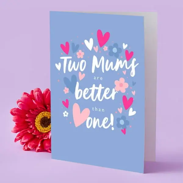 A light blue Mother’s Day card decorated with pink and blue hearts and flowers, placed upright beside a bright pink flower.