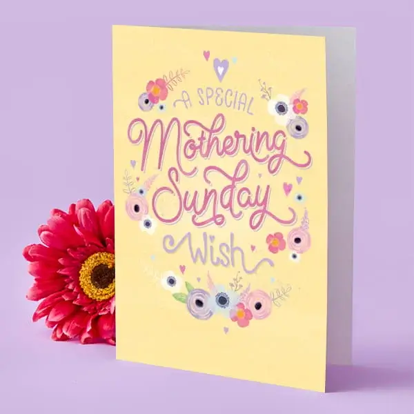 A yellow Mother’s Day card decorated with pastel flowers and hearts, displayed upright beside a bright pink flower.