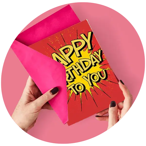 Hands holding a bold comic‑style Happy Birthday card with a bright pink envelope against a pink background.