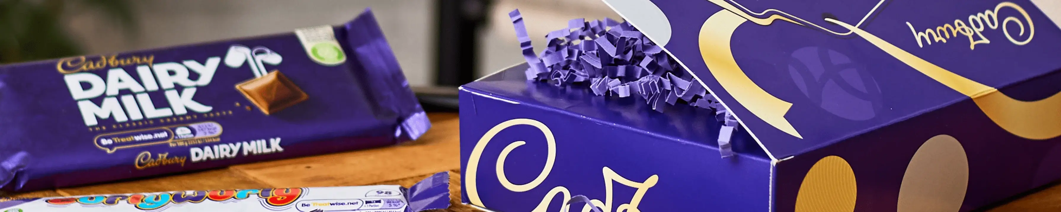 The image shows a Cadbury Dairy Milk chocolate bar and a partially opened purple gift box with the word 'Cadbury' written on it in white. The box is filled with purple shredded paper.' The image shows a Cadbury Dairy Milk chocolate bar and a partially opened purple gift box with the word 'Cadbury' written on it in white. The box is filled with purple shredded paper.'