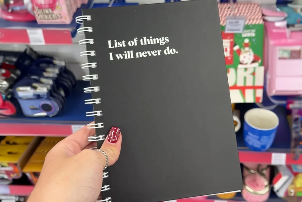 A hand holding a black spiral notebook with white text on the cover that reads “List of things I will never do,” displayed in a store aisle with other colorful items in the background. A hand holding a black spiral notebook with white text on the cover that reads “List of things I will never do,” displayed in a store aisle with other colorful items in the background.