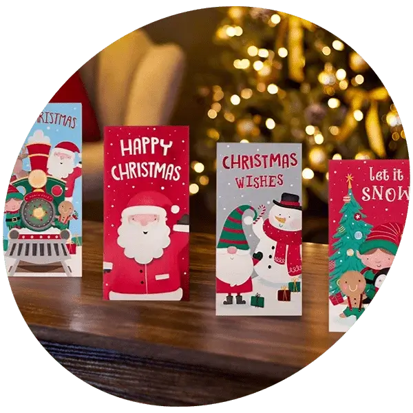 Four festive Christmas cards displayed on a wooden surface in front of a blurred Christmas tree with lights. Designs include Santa, a snowman, a train, and a tree with text such as Happy Christmas, Christmas Wishes, and Let it Snow.