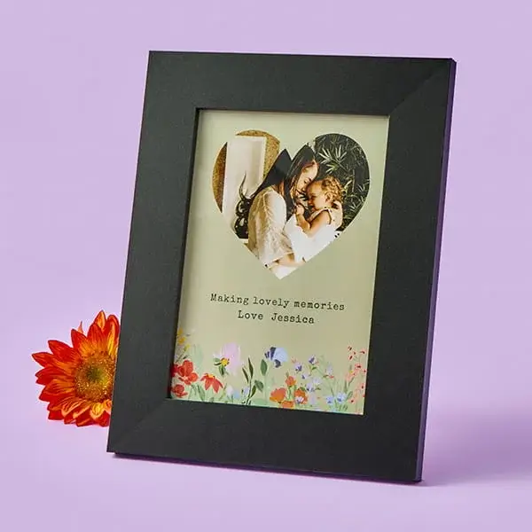 Black photo frame displaying a heart‑shaped picture of a parent and child, with floral artwork and a personalised message below, placed beside an orange flower on a pastel purple background.