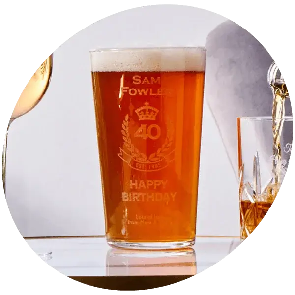 Personalised engraved pint glass filled with beer, displayed alongside champagne flutes against a light background.