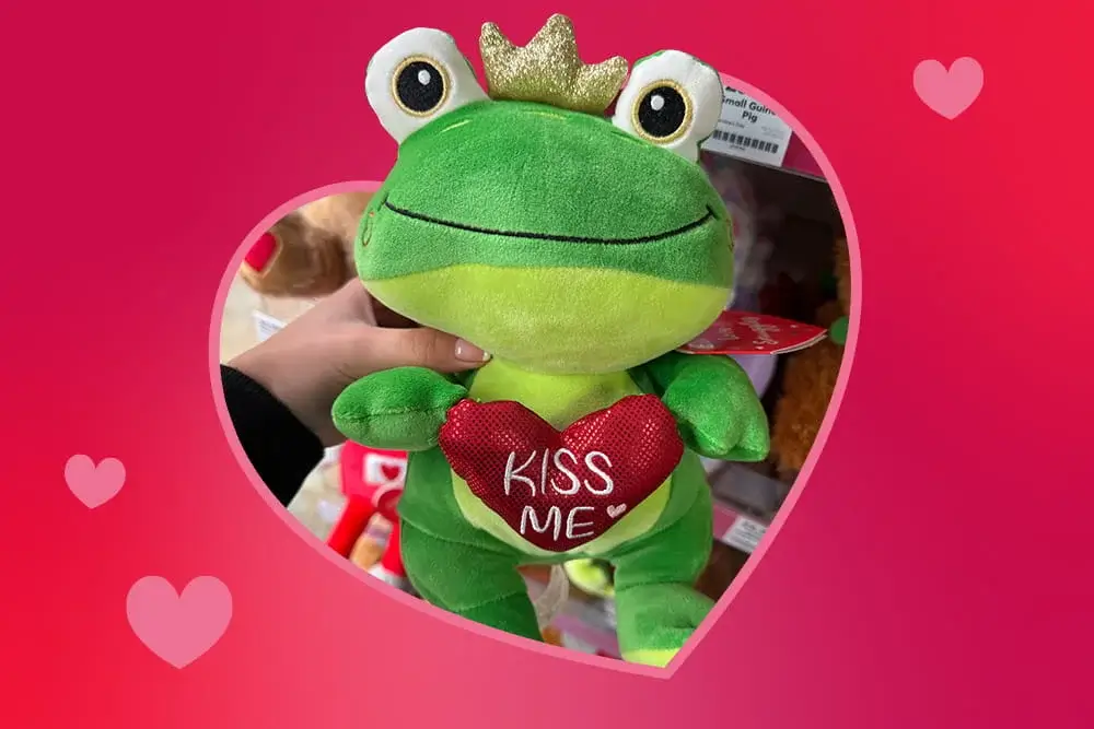 Green plush frog with a gold crown holding a red heart that says kiss me, shown against a pink heart‑themed background.