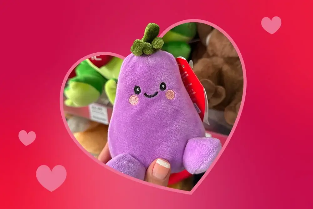 Cute purple plush toy shaped like an eggplant with a smiling face, held up in front of a shelf of stuffed animals.