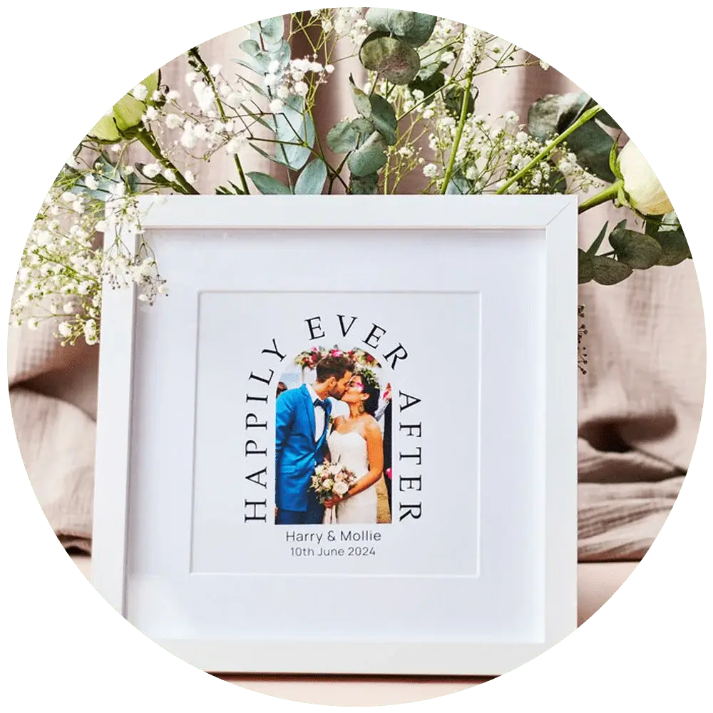 A framed photo of a couple on their wedding day. The frame is white and square, with the words 'Happily Ever After' curved above the photo. Below the photo, it says 'Harry & Mollie 10th June 2024'. The frame is surrounded by delicate white flowers and greenery. A framed photo of a couple on their wedding day. The frame is white and square, with the words 'Happily Ever After' curved above the photo. Below the photo, it says 'Harry & Mollie 10th June 2024'. The frame is surrounded by delicate white flowers and greenery.