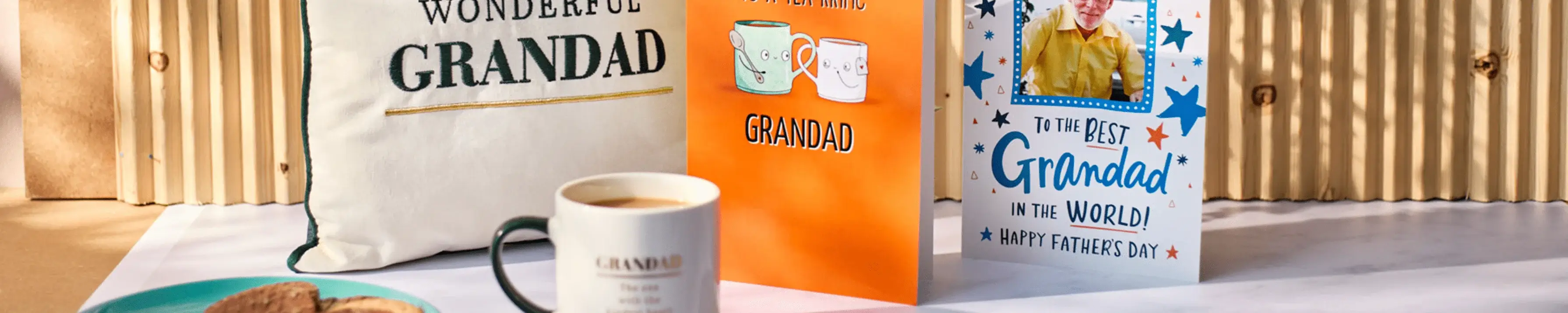 A scene featuring a 'Wonderful Grandad' cushion, an orange card with illustrations of mugs and the text 'For a Wonderful Grandad', another card with the text 'To the Best Grandad in the World! Happy Father's Day' and a photo of an elderly man, a cup of coffee labeled 'Grandad', and some biscuits on a table.' A scene featuring a 'Wonderful Grandad' cushion, an orange card with illustrations of mugs and the text 'For a Wonderful Grandad', another card with the text 'To the Best Grandad in the World! Happy Father's Day' and a photo of an elderly man, a cup of coffee labeled 'Grandad', and some biscuits on a table.'