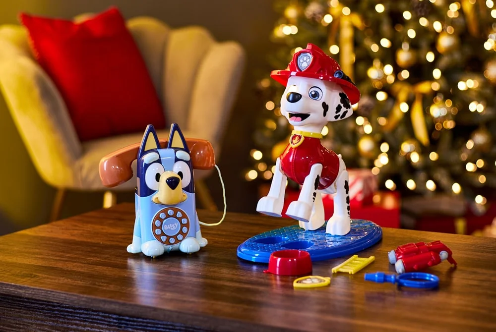 Two children’s toys, a Bluey-themed toy phone and a Paw Patrol firefighter dog figure with accessories, displayed on a wooden table in front of a decorated Christmas tree and festive lights. Two children’s toys, a Bluey-themed toy phone and a Paw Patrol firefighter dog figure with accessories, displayed on a wooden table in front of a decorated Christmas tree and festive lights.