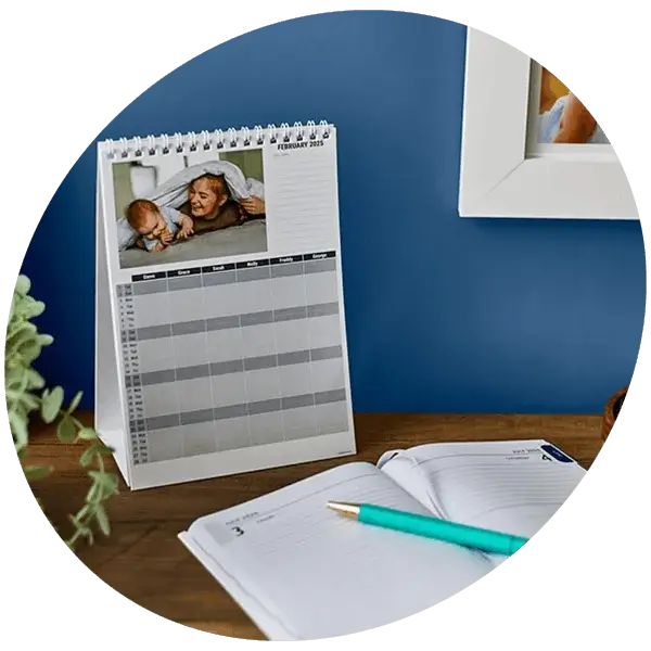 Personalised photo desk calendar with monthly planner layout displayed on a wooden desk alongside an open diary and pen.