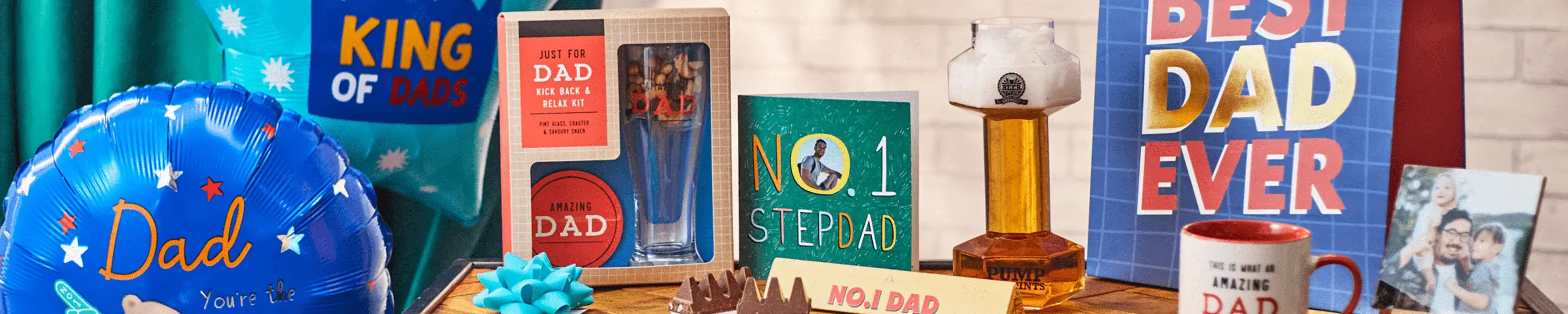 A collection of Father's Day gifts displayed on a table. Items include a blue balloon with 'Dad' written on it, a 'King of Dads' sign, a boxed set containing a glass and coaster labeled 'Dad', various greeting cards including one that says 'No. 1 Stepdad', and another that says 'Best Dad Ever'. There is also a bottle of amber liquid, a mug with the word 'Dad' on it, and other small gift items.' A collection of Father's Day gifts displayed on a table. Items include a blue balloon with 'Dad' written on it, a 'King of Dads' sign, a boxed set containing a glass and coaster labeled 'Dad', various greeting cards including one that says 'No. 1 Stepdad', and another that says 'Best Dad Ever'. There is also a bottle of amber liquid, a mug with the word 'Dad' on it, and other small gift items.'