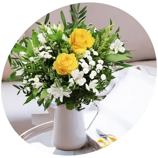 Bouquet with yellow roses, small white flowers, and green foliage arranged in a white pitcher.