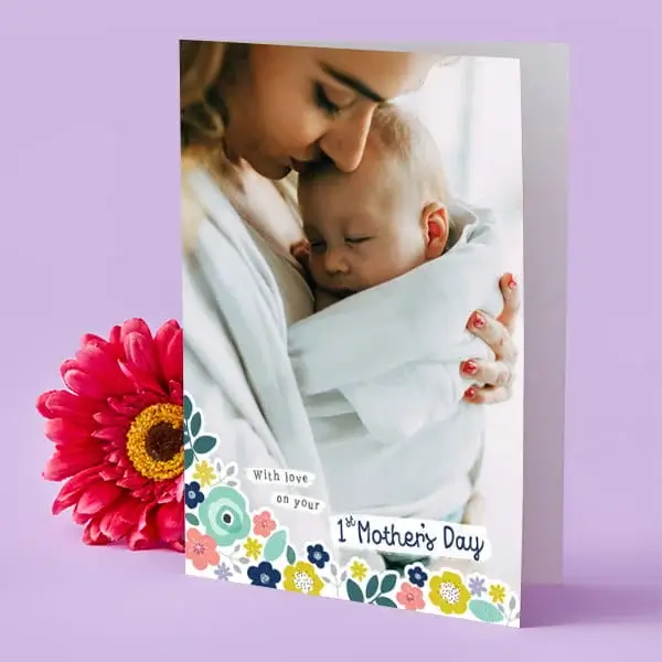 A floral Mother’s Day card standing upright, featuring an adult gently holding a baby, with a bright pink flower placed beside it on a purple background.